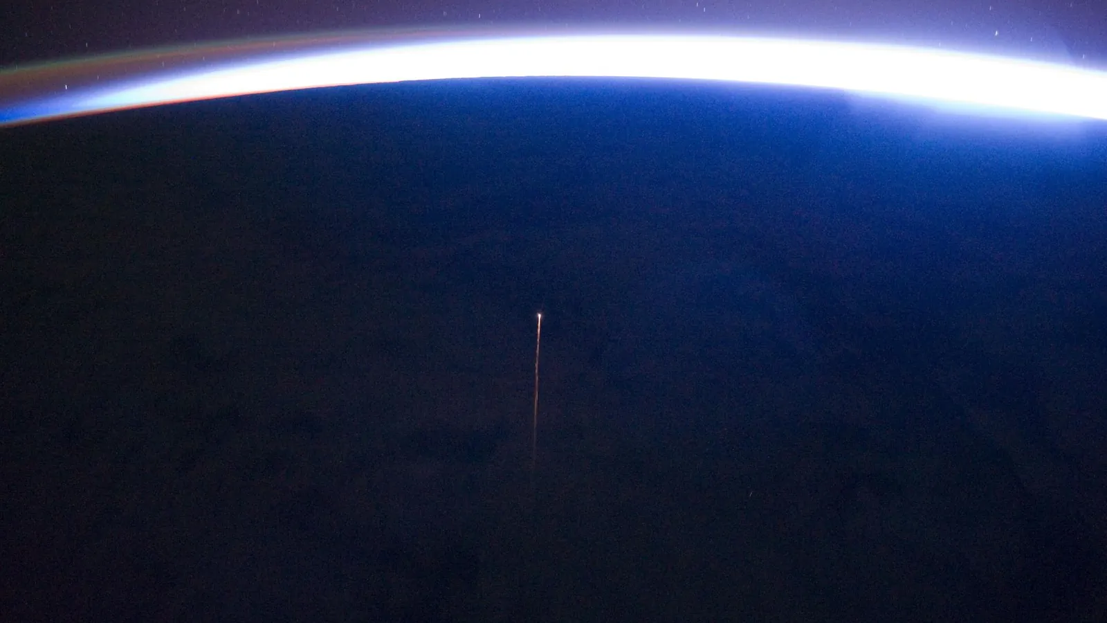 从国际空间站俯拍的地球夜面，地平线上方是明亮的大气辉光，下方一道橙色细线是进步号货运飞船再入时的等离子体尾迹